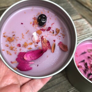 Pu&ograve; includere: Una candela viola con petali di fiori secchi e una pietra nera in cima. La candela &egrave; in una latta d'argento.
