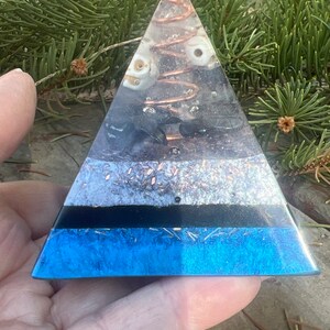 May include: A clear pyramid made of resin with layers of different crystals and a copper coil inside. The pyramid is blue at the base and has a black layer above it. The pyramid is sitting on a hand.