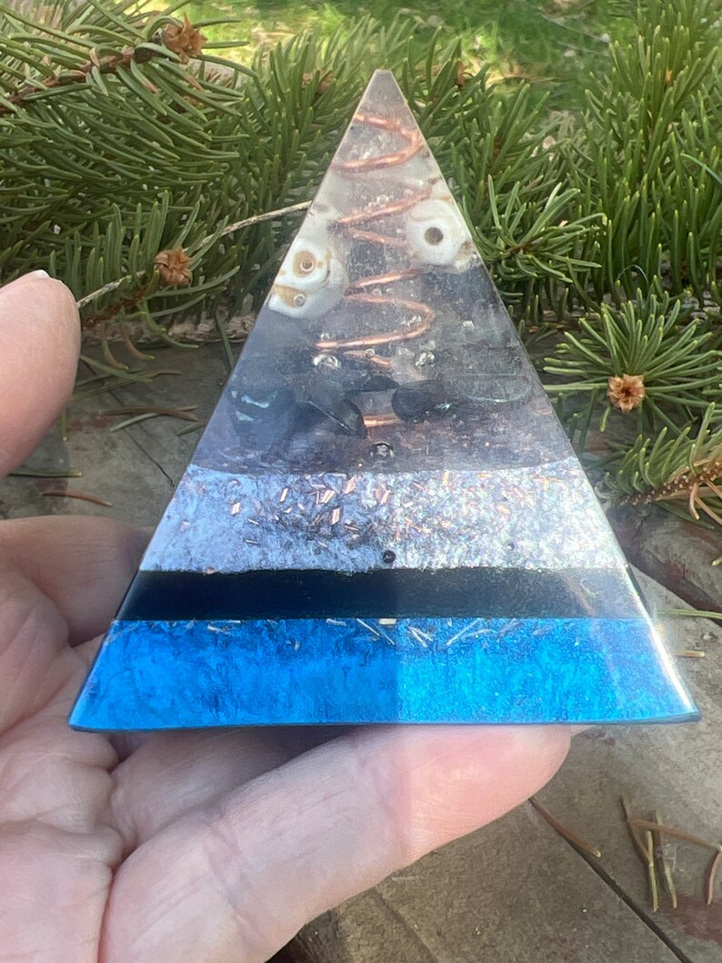 May include: A clear pyramid made of resin with layers of different crystals and a copper coil inside. The pyramid is blue at the base and has a black layer above it. The pyramid is sitting on a hand.
