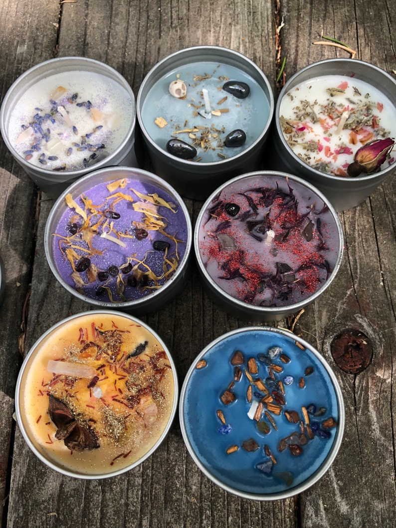 Pu&ograve; includere: Sette piccole lattine di candele rotonde di colore argento con diverse cere colorate e cristalli, erbe e altri elementi naturali incorporati. Le candele sono disposte in cerchio su una superficie di legno.