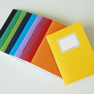 Puede incluir: Una colección de cuadernos coloridos en varios tonos, incluyendo negro, verde, azul, morado, naranja y amarillo. Los cuadernos tienen un diseño sencillo con una etiqueta en blanco en la portada del cuaderno amarillo. Los cuadernos están dispuestos en forma de abanico.