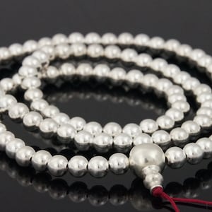 May include: A silver beaded mala necklace with a large, rounded bead at the end and a red tassel. The necklace is composed of many small, round beads. The beads are reflective and appear to be made of metal.