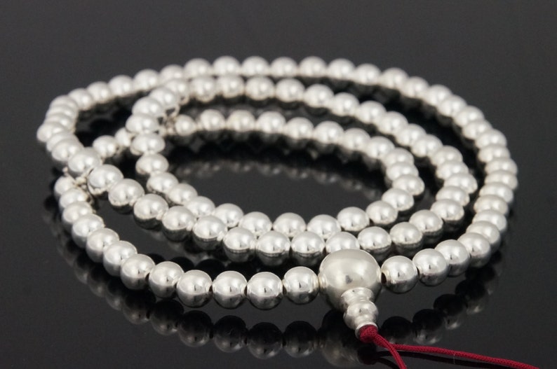 May include: A silver beaded mala necklace with a large, rounded bead at the end and a red tassel. The necklace is composed of many small, round beads. The beads are reflective and appear to be made of metal.