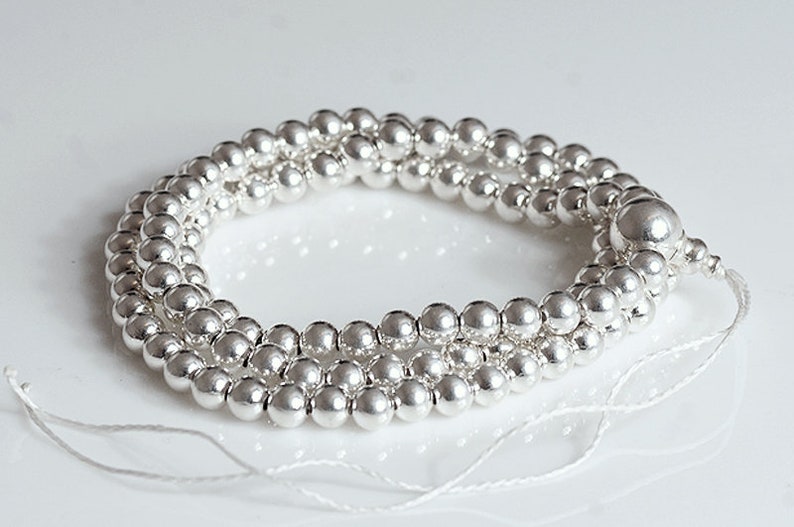 May include: A strand of shiny, silver-colored beads. The beads are small and round, strung together on a white string. The beads are arranged in a circular shape, with the ends of the string visible. The beads are reflective and appear to be made of metal.