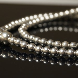 May include: A close-up of a silver beaded necklace. The necklace is composed of multiple strands of small, round, shiny beads. The beads reflect light, creating a bright, metallic appearance. A red string and white tag are attached.