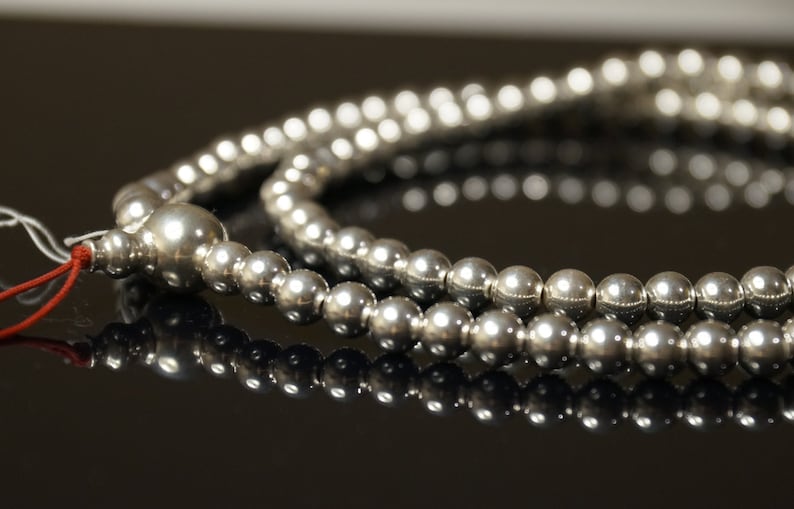 May include: A close-up of a silver beaded necklace. The necklace is composed of multiple strands of small, round, shiny beads. The beads reflect light, creating a bright, metallic appearance. A red string and white tag are attached.