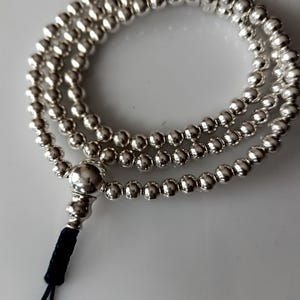 May include: A circular strand of polished silver-coloured beads arranged in three concentric loops. A larger, smooth silver-coloured bead connects to a decorative silver-coloured component, which is attached to a thin black cord loop at the end.