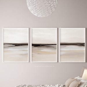 May include: Three abstract landscape paintings in white frames, featuring a muted color palette of beige, brown, and white. The paintings are hung on a light pink wall above a bed with a white linen duvet and a woven rug.
