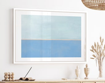 Op de afbeelding: Een ingelijst abstract schilderij dat een blauwe en lichtblauwe lucht met een dunne lijn van beige zand aan de onderkant toont. Het schilderij hangt aan een witte muur boven een houten kast met geweven rotan deuren.