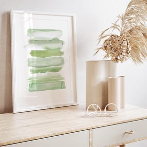 May include: A framed abstract watercolor painting with green brushstrokes in a white frame on a wooden desk with two drawers. A vase with dried flowers and a pair of round glasses are on the desk.