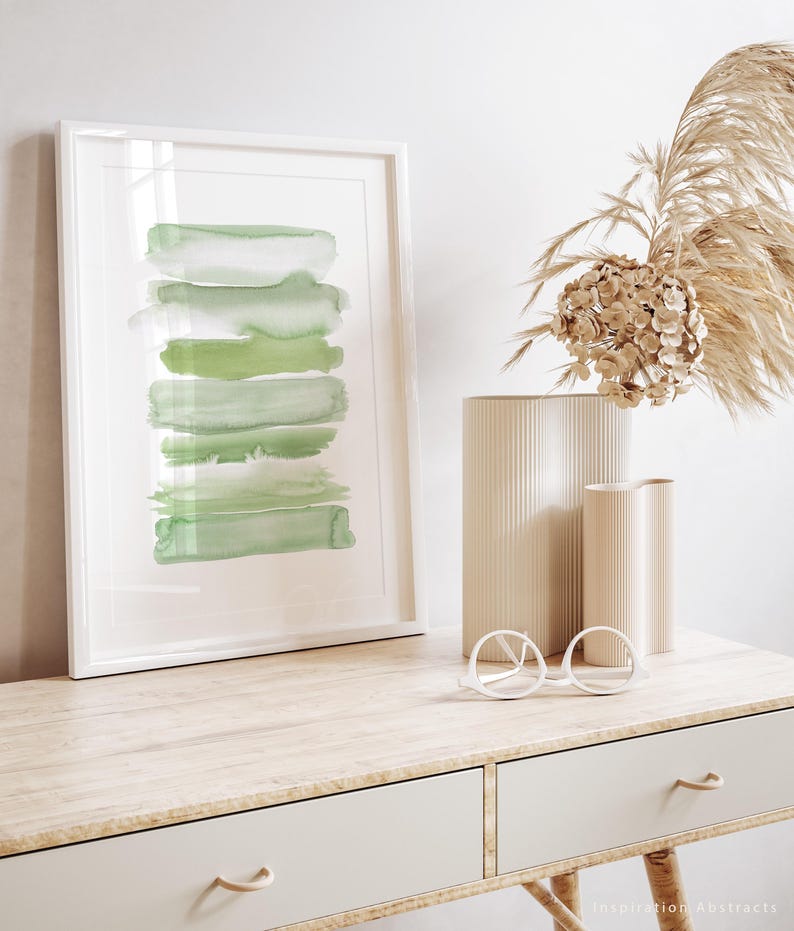 May include: A framed abstract watercolor painting with green brushstrokes in a white frame on a wooden desk with two drawers. A vase with dried flowers and a pair of round glasses are on the desk.