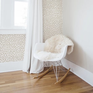May include: A modern white rocking chair with a fluffy, cream-colored throw. The chair has a metal frame and wooden rockers. The background features a wall with a beige, spotted pattern, a window, and a white curtain.