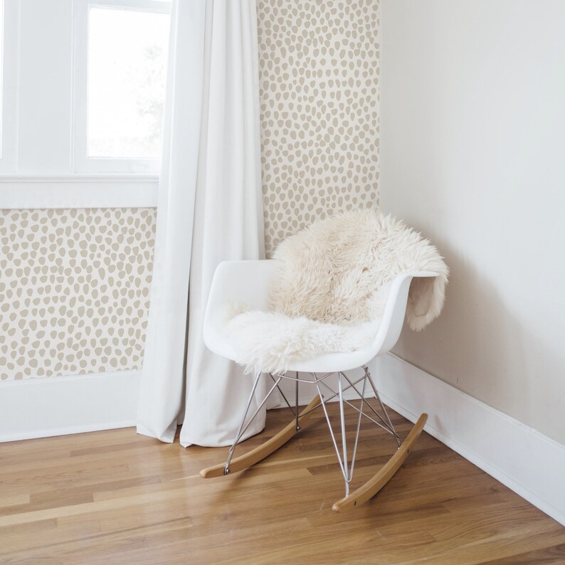 May include: A modern white rocking chair with a fluffy, cream-colored throw. The chair has a metal frame and wooden rockers. The background features a wall with a beige, spotted pattern, a window, and a white curtain.