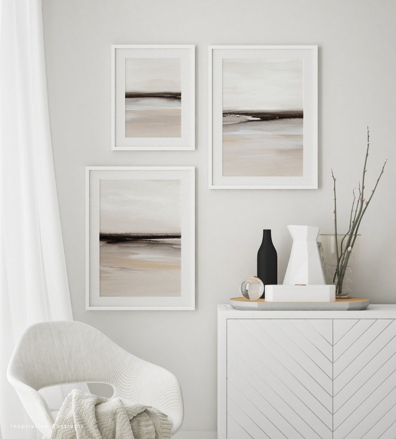 May include: Three framed abstract landscape paintings in muted tones of brown, beige, and white, hanging on a white wall above a white dresser with a chevron pattern.
