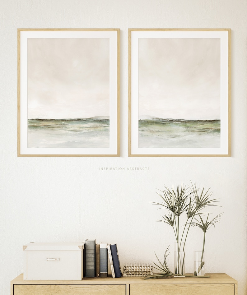 May include: Two framed abstract landscape paintings with a muted color palette of beige, green, and white. The paintings are hung on a white wall above a wooden shelf with a white box, books, and a vase of green plants.