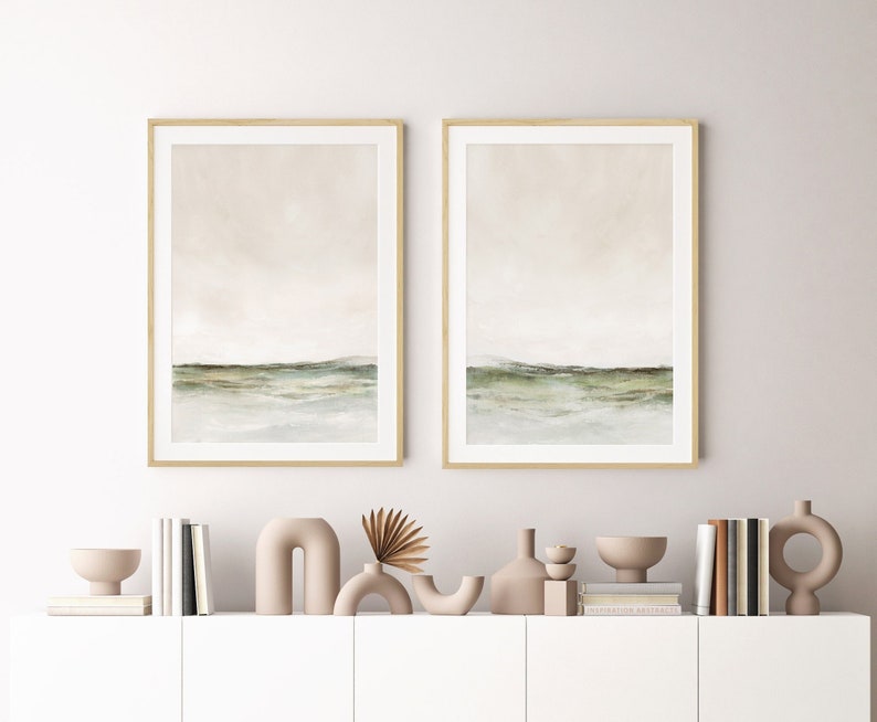 May include: Two framed watercolor paintings of a landscape with a green hill and a light blue sky. The paintings are hanging on a white wall above a white shelf with decorative objects.