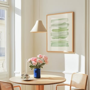 May include: A light wood dining table with two chairs, a vase of pink flowers, and a white pendant light hanging above. The table is set with a white plate and a blue vase.