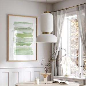May include: A white pendant light with a textured finish hangs from the ceiling above a wooden table. The light has a gold trim and a white shade. A framed abstract painting with green watercolor stripes hangs on the wall behind the table.