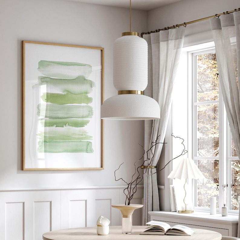 May include: A white pendant light with a textured finish hangs from the ceiling above a wooden table. The light has a gold trim and a white shade. A framed abstract painting with green watercolor stripes hangs on the wall behind the table.