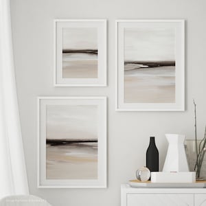 May include: Three framed abstract landscape paintings in muted tones of brown, beige, and white. The paintings are hung on a white wall in a modern interior setting.