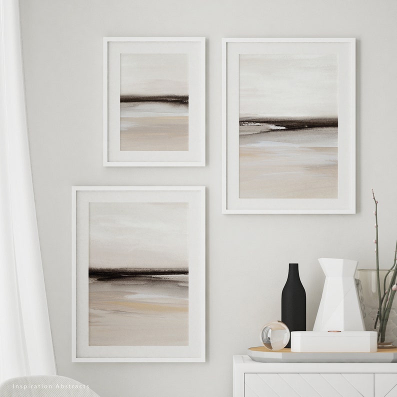 May include: Three framed abstract landscape paintings in muted tones of brown, beige, and white. The paintings are hung on a white wall in a modern interior setting.