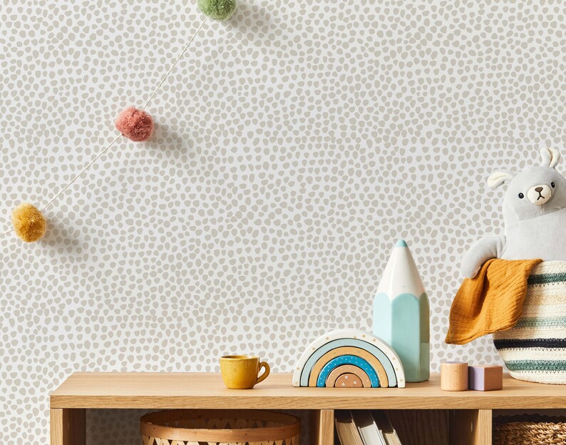 May include: A wooden shelf displays children's decor against a speckled wallpaper backdrop. Items include a yellow mug, a rainbow-shaped wooden toy, a blue and white pencil-shaped decoration, and a gray stuffed animal in a woven basket. A garland of colorful pom-poms hangs above.