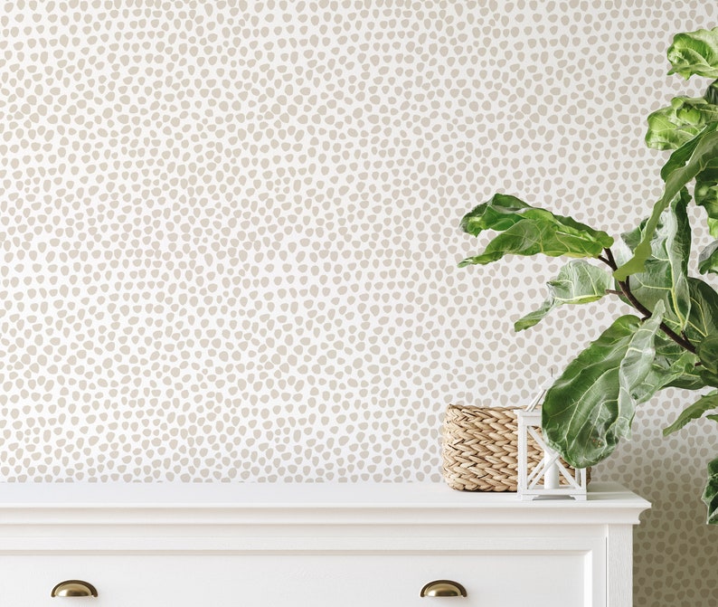 May include: Neutral-toned wallpaper with a speckled pattern, creating a subtle, textured backdrop. A white dresser with brass hardware and a woven basket add to the room's decor. A large potted plant with green leaves is on the right.