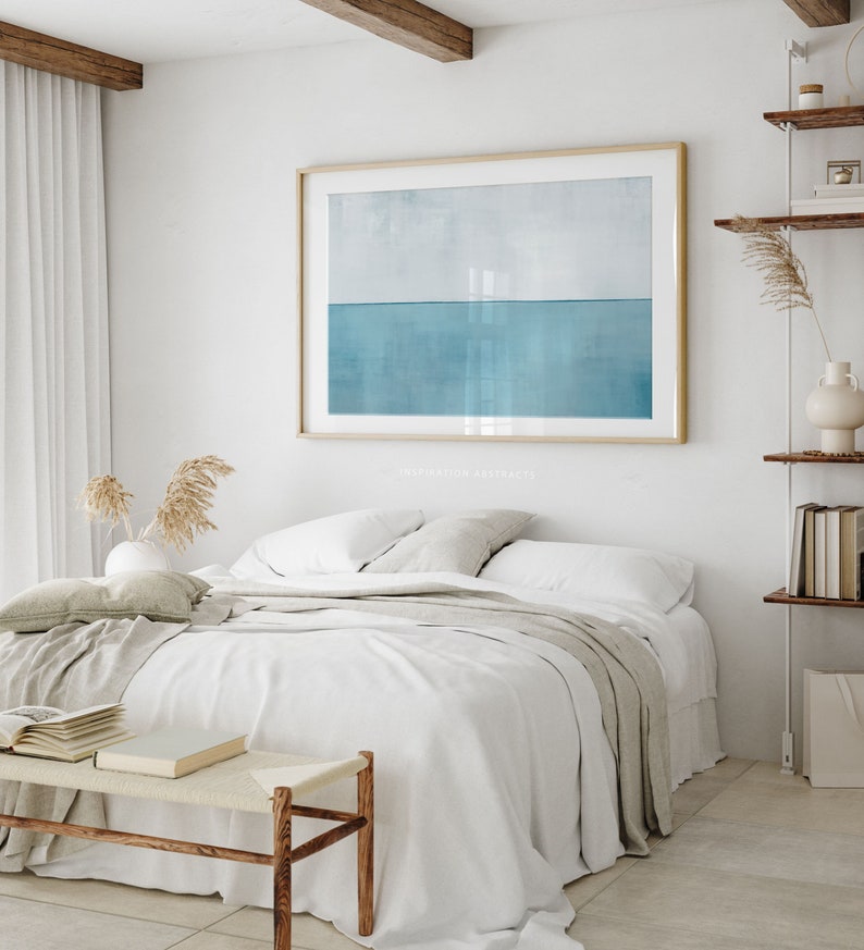 May include: A white bed with a light gray throw blanket and two pillows. A wooden bench with a woven seat sits in front of the bed. A framed abstract painting of a blue and white horizon hangs on the wall above the bed.