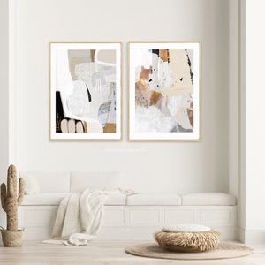 May include: Two abstract art prints in beige, brown, and white tones with a wood frame. The prints are hung on a white wall above a white bench with a beige throw blanket and a woven ottoman.
