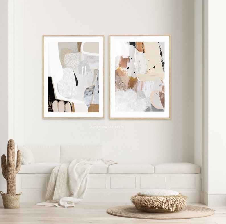 May include: Two abstract art prints in beige, brown, and white tones with a wood frame. The prints are hung on a white wall above a white bench with a beige throw blanket and a woven ottoman.
