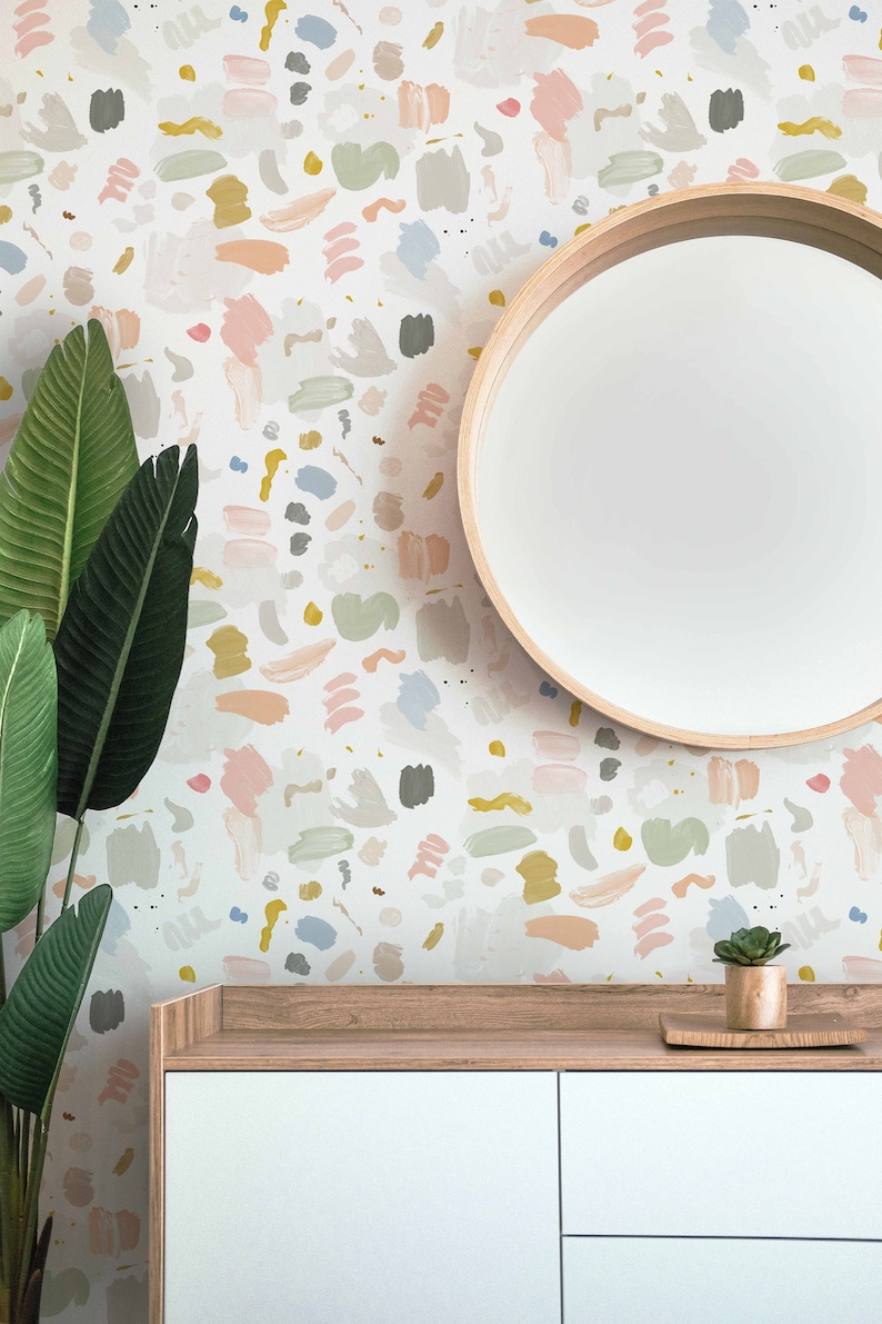 May include: A white wall with a colorful abstract pattern wallpaper. A round wooden mirror hangs on the wall above a white dresser with wooden top. A small potted succulent plant sits on the dresser.