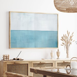 May include: A framed abstract painting with a light blue and white color scheme. The painting is hanging on a white wall above a wooden cabinet with woven rattan doors.