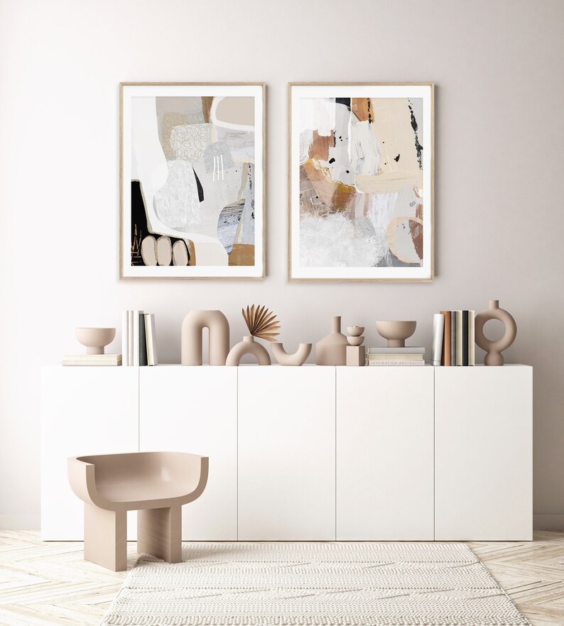May include: Two abstract art prints in beige, brown, and white frames, hanging above a white cabinet with beige ceramic sculptures and books.