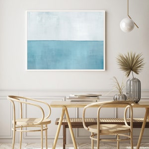 May include: A light blue abstract painting with a horizontal line dividing the canvas into two sections. The top section is a lighter shade of blue, while the bottom section is a darker shade of blue. The painting is framed in a white frame and is hanging on a white wall. The painting is in a room with a wooden table and chairs.