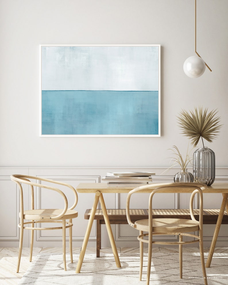 May include: A light blue abstract painting with a horizontal line dividing the canvas into two sections. The top section is a lighter shade of blue, while the bottom section is a darker shade of blue. The painting is framed in a white frame and is hanging on a white wall. The painting is in a room with a wooden table and chairs.