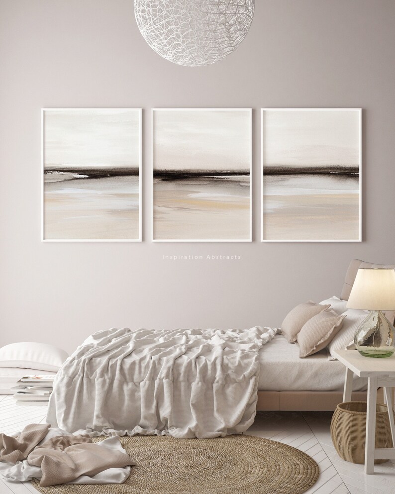 May include: Three abstract landscape paintings in white frames, featuring a muted color palette of beige, brown, and white. The paintings are hung on a light pink wall above a bed with a white linen duvet and a woven rug.