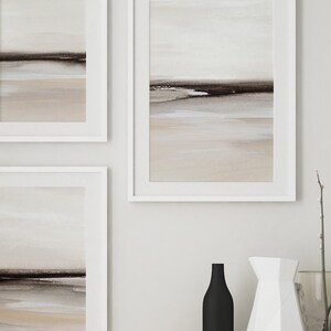 May include: Three framed abstract landscape paintings in muted tones of brown, beige, and white, hanging on a white wall. The paintings are in white frames and feature a horizontal line of dark brown paint across the middle of each canvas, representing a horizon line.