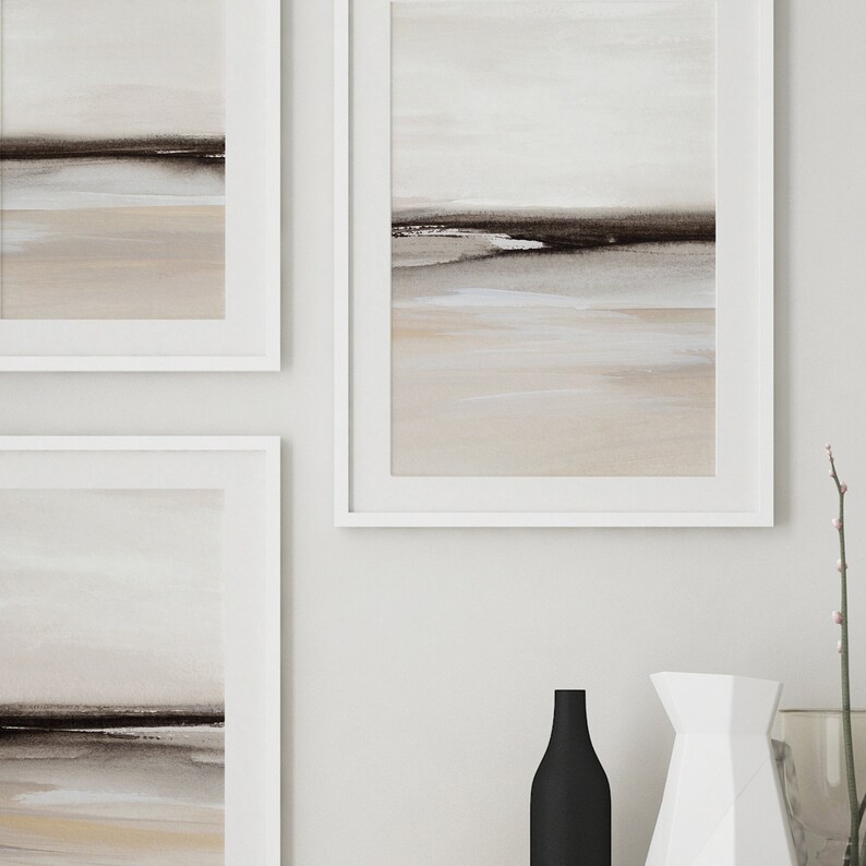 May include: Three framed abstract landscape paintings in muted tones of brown, beige, and white, hanging on a white wall. The paintings are in white frames and feature a horizontal line of dark brown paint across the middle of each canvas, representing a horizon line.
