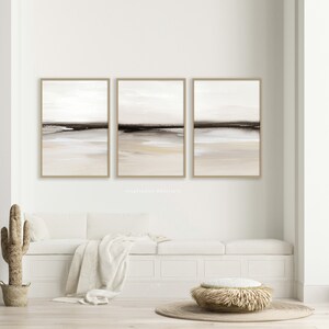 May include: Three abstract landscape paintings in muted tones of brown, beige, and white. The paintings are framed in a light brown wood frame and are hung on a white wall.