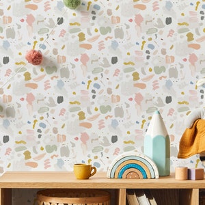 May include: A white wall with a colorful abstract pattern wallpaper. The wallpaper features a repeating pattern of brushstrokes in shades of pink, blue, gray, and yellow. The wall is decorated with a string of pom-poms, a rainbow stacking toy, a blue pencil, and a stuffed animal.