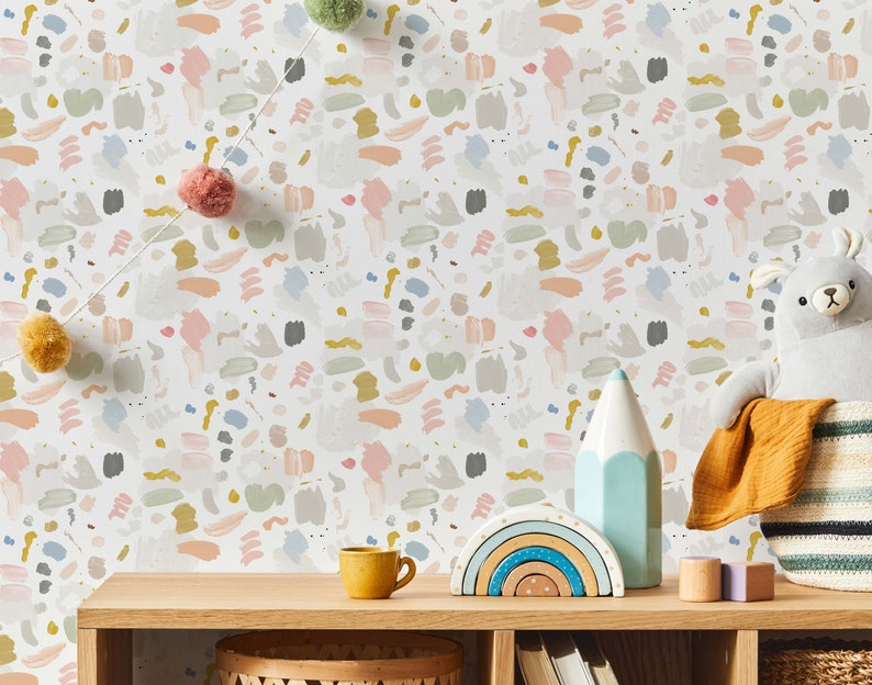 May include: A white wall with a colorful abstract pattern wallpaper. The wallpaper features a repeating pattern of brushstrokes in shades of pink, blue, gray, and yellow. The wall is decorated with a string of pom-poms, a rainbow stacking toy, a blue pencil, and a stuffed animal.
