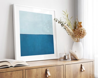 Op de afbeelding: Een ingelijste abstracte kunstprint met een lichtblauwe bovenhelft en een donkerblauwe onderhelft. De print hangt aan een witte muur boven een houten kast met twee laden. Een vaas met gedroogde bloemen staat op de kast.