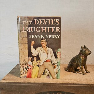 Puede incluir: Un libro de bolsillo vintage titulado "The Devil's Laughter" de Frank Yerby. La portada muestra a un hombre con una camisa blanca y pantalones marrones, con un grupo de personas en el fondo. Una figura de bulldog de bronce se sienta sobre una caja de madera con el texto "Cal Pack From The Land Of Sunshine Fruits" impreso en ella.