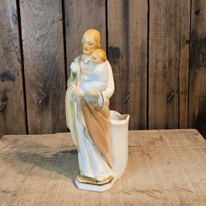 Könnte beinhalten: Keramikfigur einer religiösen Figur, die ein Kind hält, mit einer vasenartigen Öffnung am Boden. Die Figur ist in weißen und beigen Gewändern mit goldenen Akzenten gekleidet. Der Hintergrund ist eine Holzoberfläche.