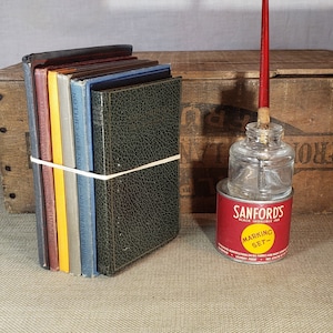 May include: A stack of vintage books with a rubber band around them, a glass bottle with a red ink pen, and a red and white label that reads "Sanford's Black Indelible Ink Marking Set -" on a wooden surface.