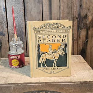 Puede incluir: Un libro antiguo titulado "The Merrill Readers Second Reader" con una ilustración colorida de una persona a caballo. Junto al libro, un tintero de cristal rojo y transparente con una pluma roja.