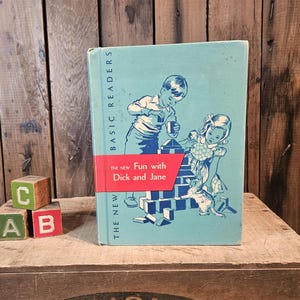 Könnte beinhalten: Ein gebundenes Vintage-Buch mit dem Titel The New Fun with Dick and Jane, das einen hellblauen Einband mit einer dunkelblauen Illustration von zwei Kindern beim Bauen mit Klötzen und einem kleinen Hund zeigt. Ein rotes Band zeigt den Titel.