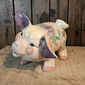 May include: A ceramic piggy bank with a pink body, purple ears, and a floral design. The flowers are painted in shades of purple, green, and yellow.