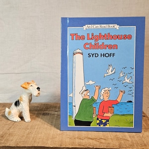 Puede incluir: Un libro infantil titulado "The Lighthouse Children" de Syd Hoff, con una cubierta azul e ilustraciones de un faro y personas. Una pequeña figura de perro de cerámica se encuentra a la izquierda sobre una superficie de madera.