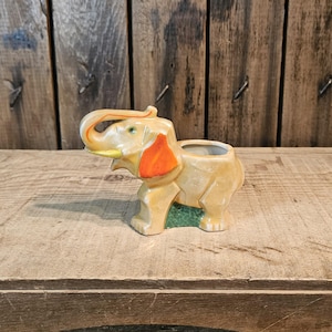 May include: A ceramic elephant planter in shades of tan, orange, and green. The elephant has a raised trunk and a small opening on its back for planting. The planter is on a wooden surface.
